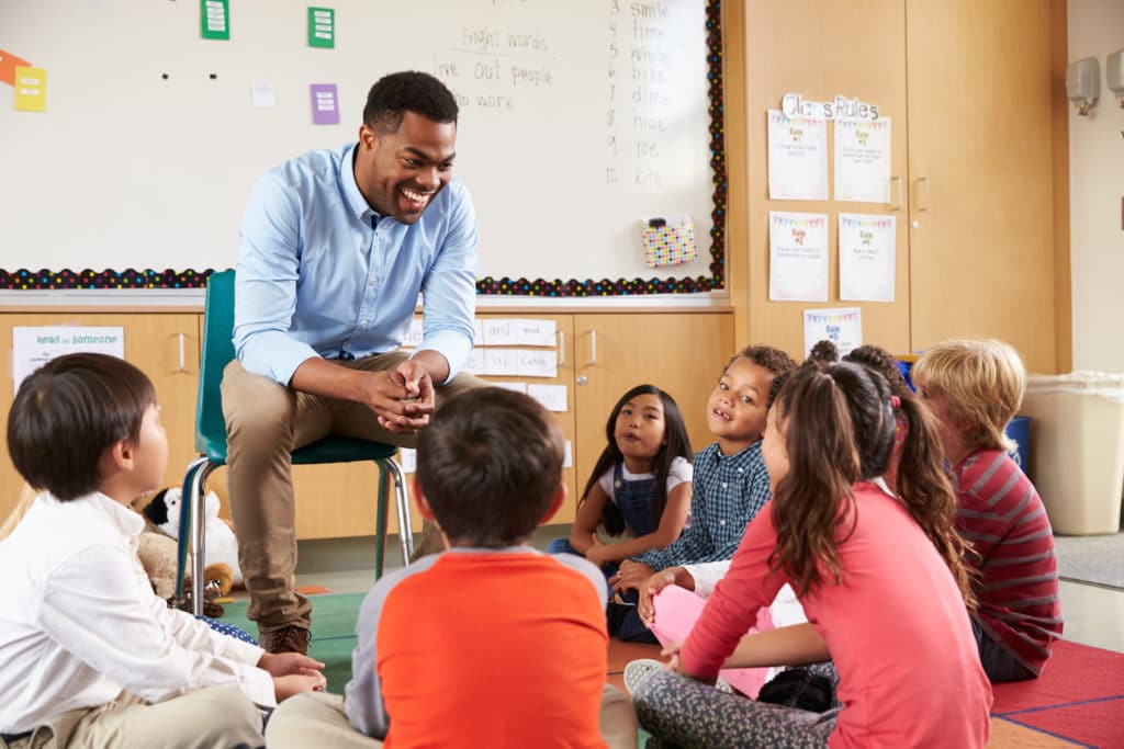 Young teacher sitting in a chair surrounded by students sitting in a circle on the floor.
