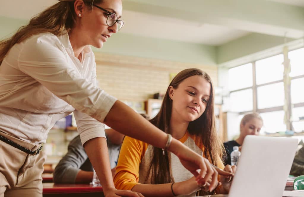 High school student working on a laptop with a teacher pointing at the screen.