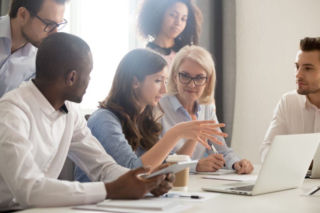 A technology mentor leads a group of professionals around a table using a computer and tablet.