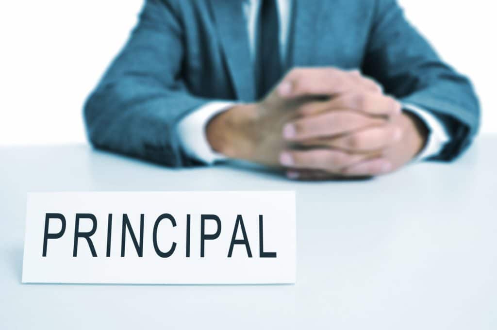 A man with a suit sits in a desk with a signboard in front of him that says “principal” on it.