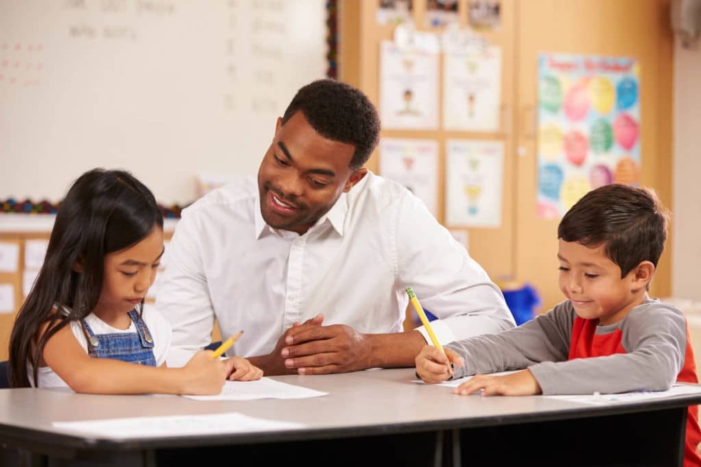 Teacher sitting with two young students writing on paper.