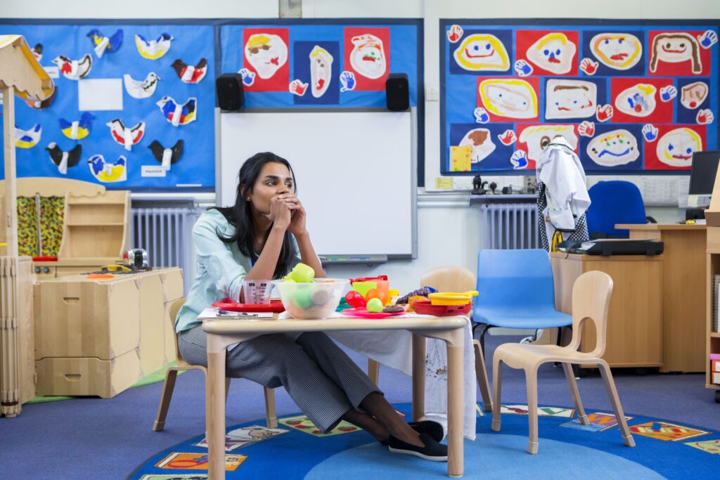 A stressed teacher sits at a table in her empty classroom.