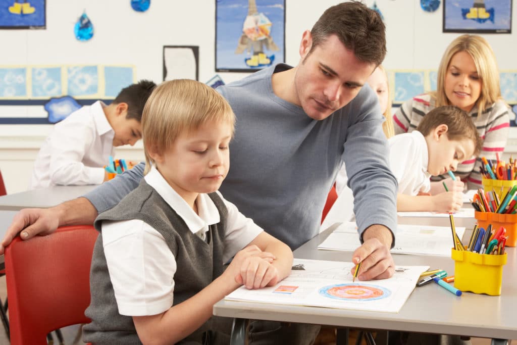 Teacher helping an uninterested student at a desk in a classroom.