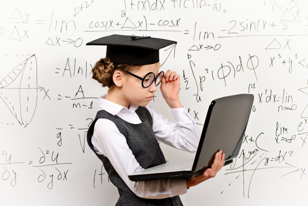 A smart schoolgirl with glasses and a graduate cap stands in front of a whiteboard with a laptop.