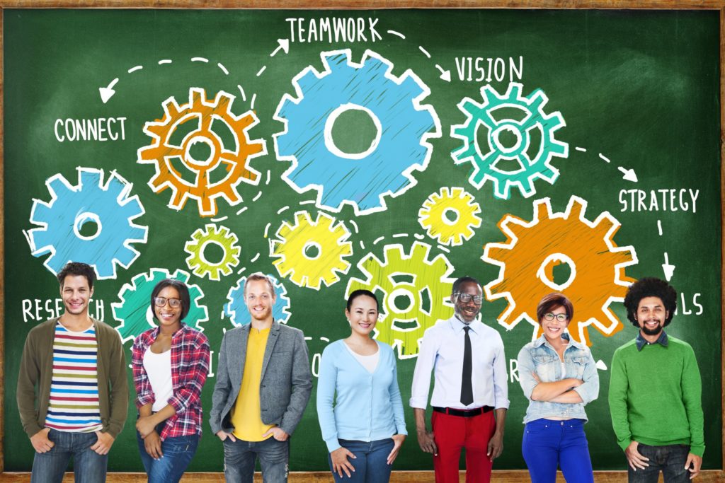 Group of educators standing in front of a chalkboard with drawings of gears and words.