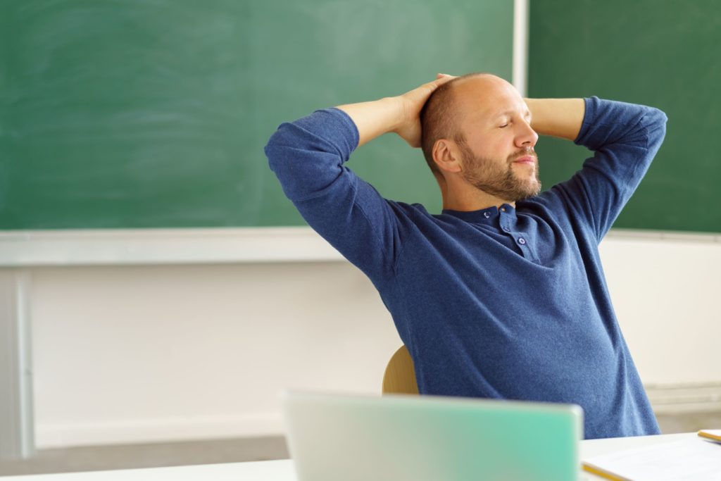 A tired teacher sits at his desk, relaxing with his eyes closed and hands behind his head.