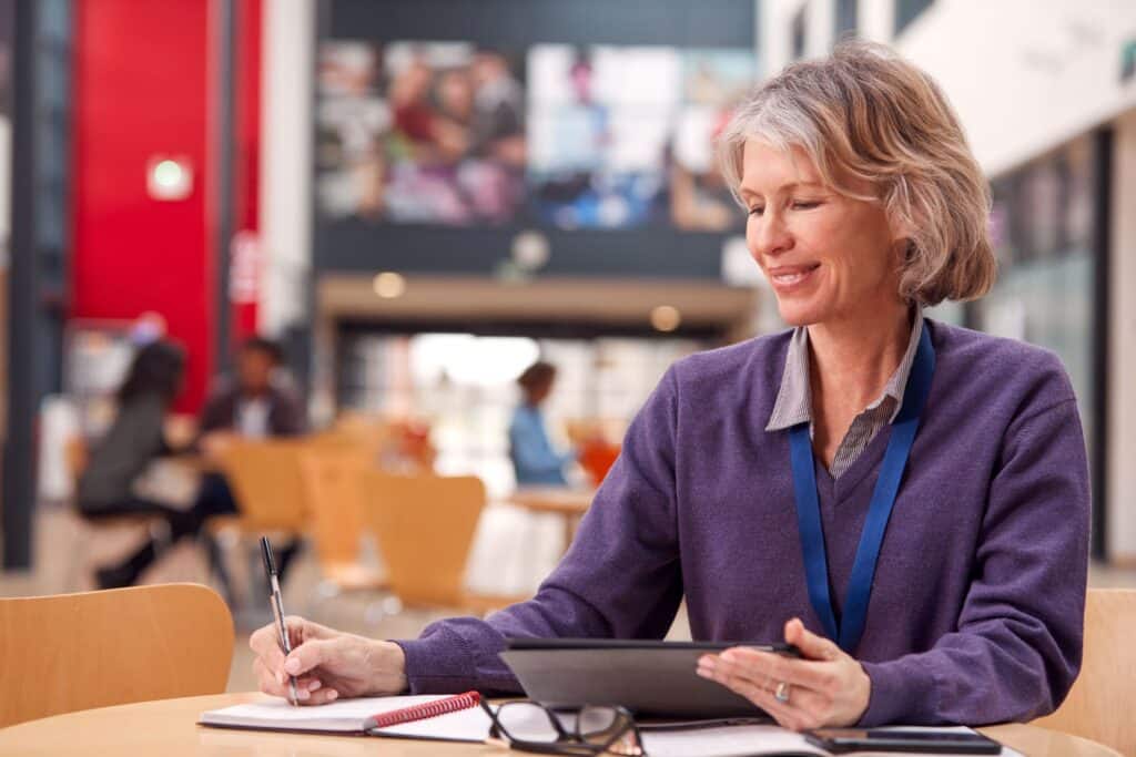A teacher sits at a table and writes something down while looking through her tablet.