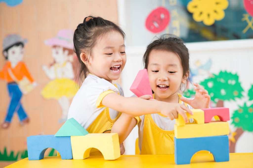 Two young girls building a block tower in a classroom.