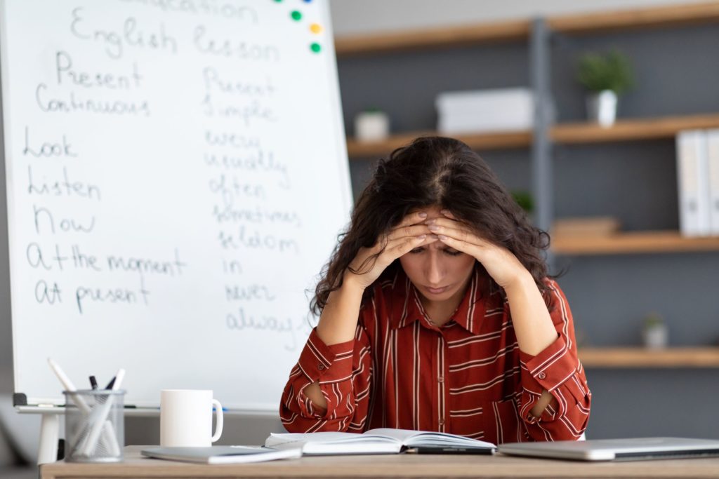 A teacher sits in the classroom at a desk, holding her head.