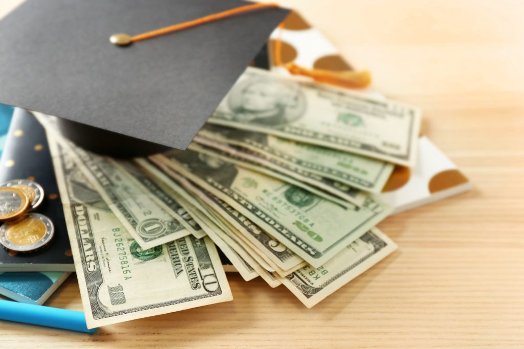School supplies, graduation hat, dollar banknotes and coins sit on a wooden table.