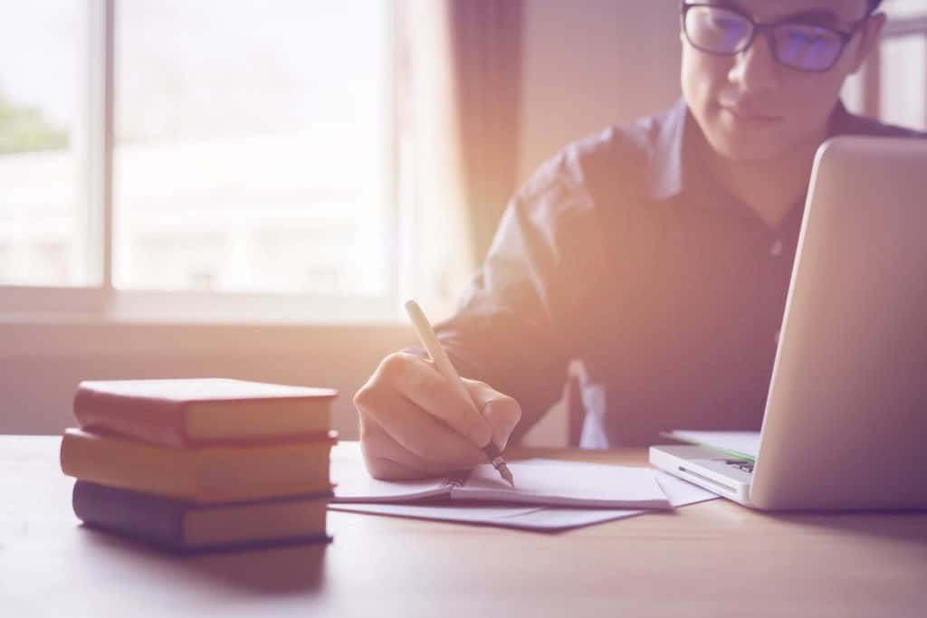 Man writes out a college application with pen and paper while looking at a school website on a laptop.