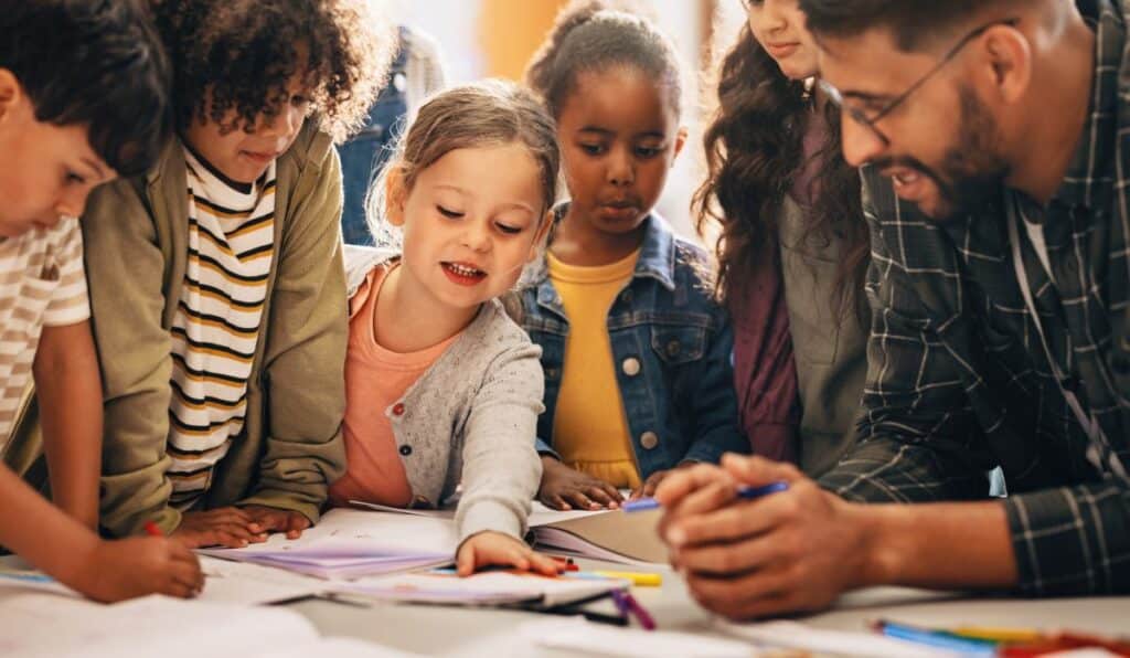 A group of primary students gather around a table with their teacher, working on an assignment.