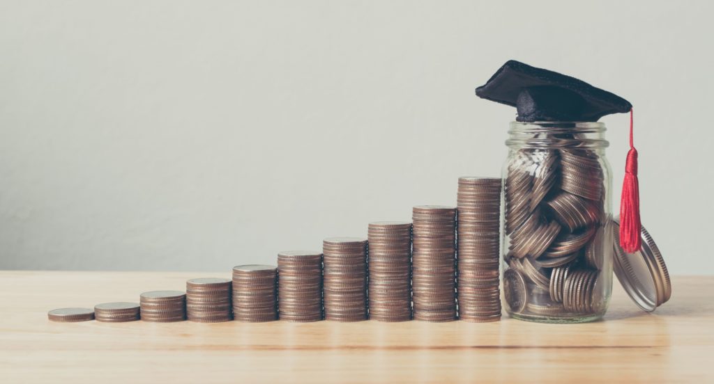 Stacks of coins in a line on a table next to a jar of coins with a graduation cap on top.