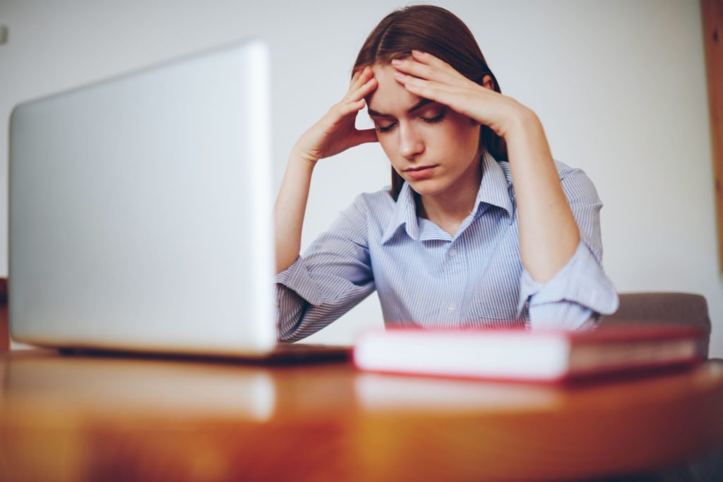 Young woman holding her head sitting at a desk with a laptop.