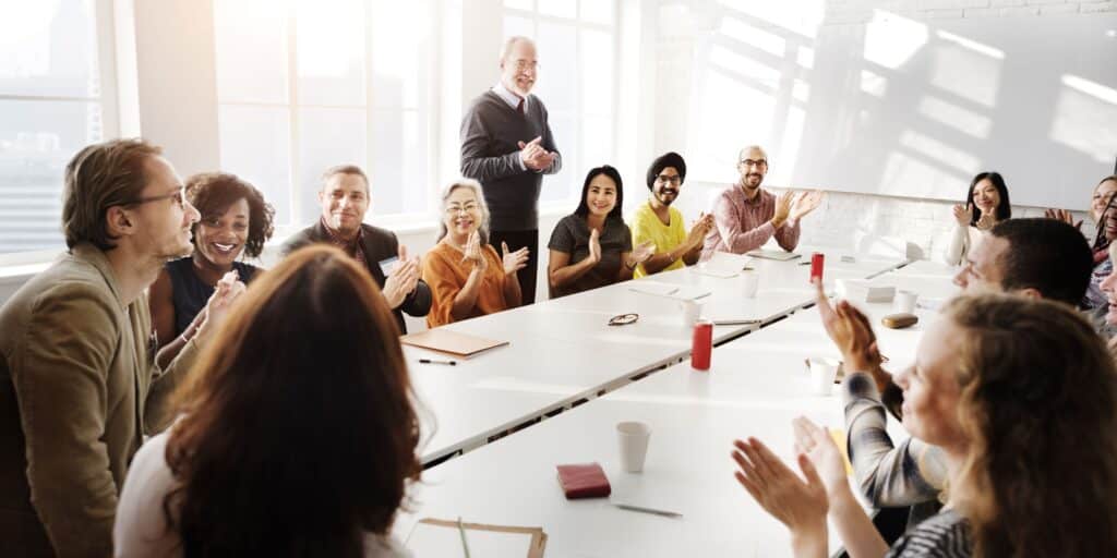 A meeting is conducted in a meeting room; all the attendees sit around a table while one stands and talks.