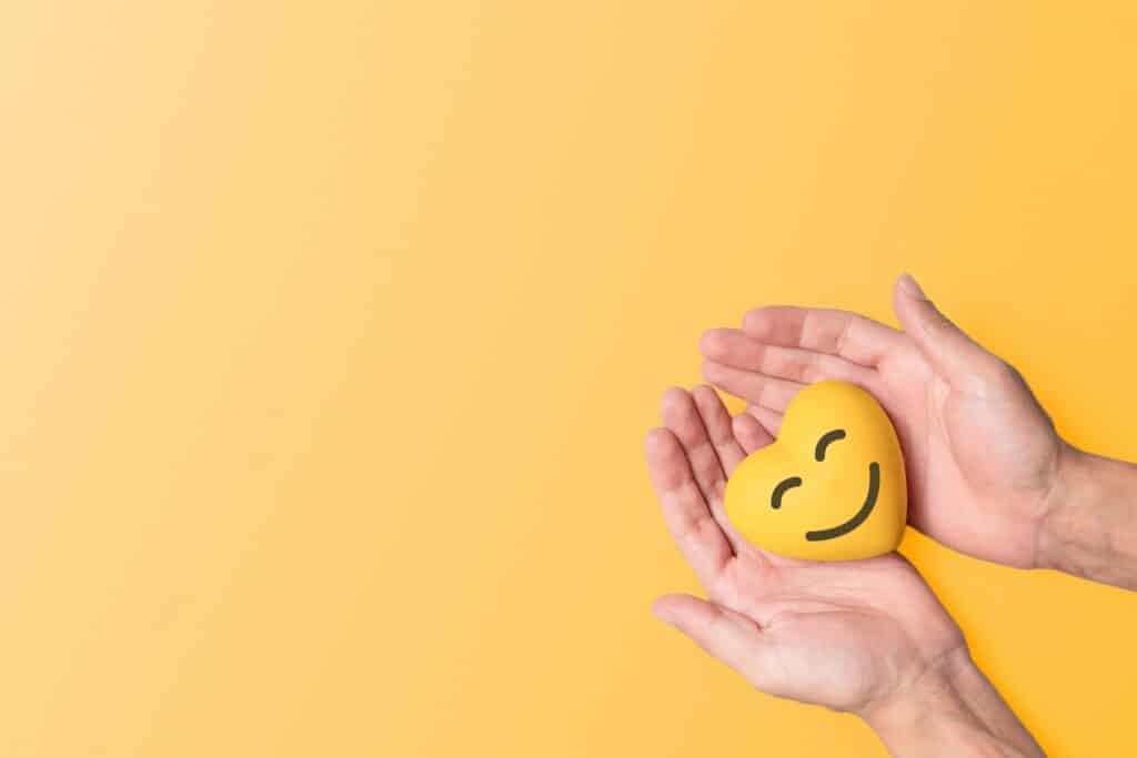 A pair of hands holds a yellow heart shaped stress ball that has a smiling face.