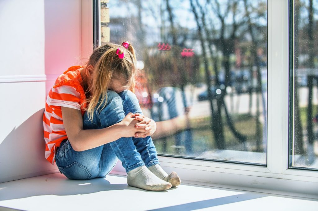 A little girl in an orange shirt sits on a window sill, crying with her face buried in her knees.