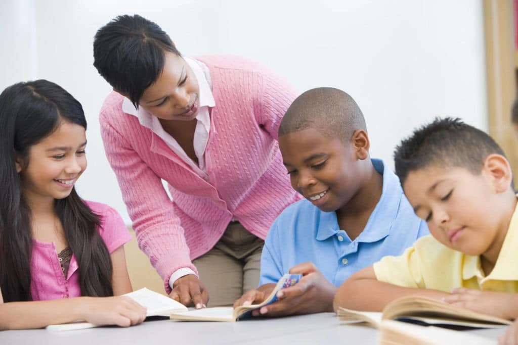 Group of young students reading at a table with a teacher checking in.
