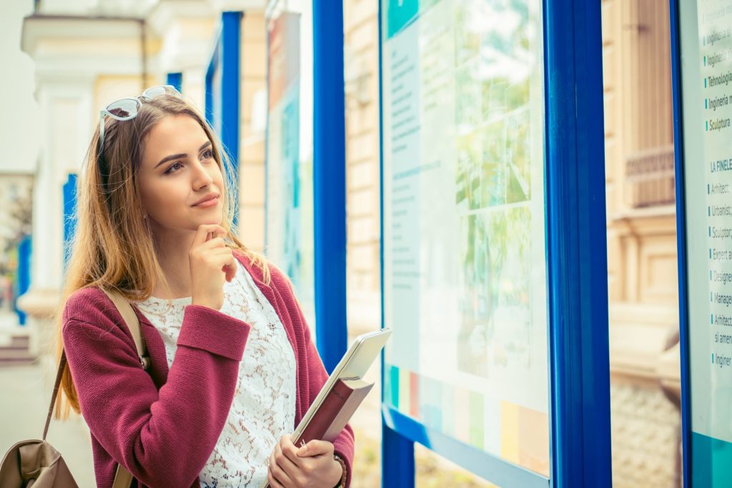 A future student stands with a book bag and books in her hands, looking at a informational college map.