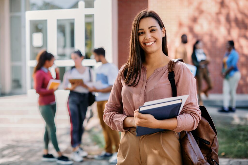 A portrait of a smiling graduate school student standing with their books in front of a university building.