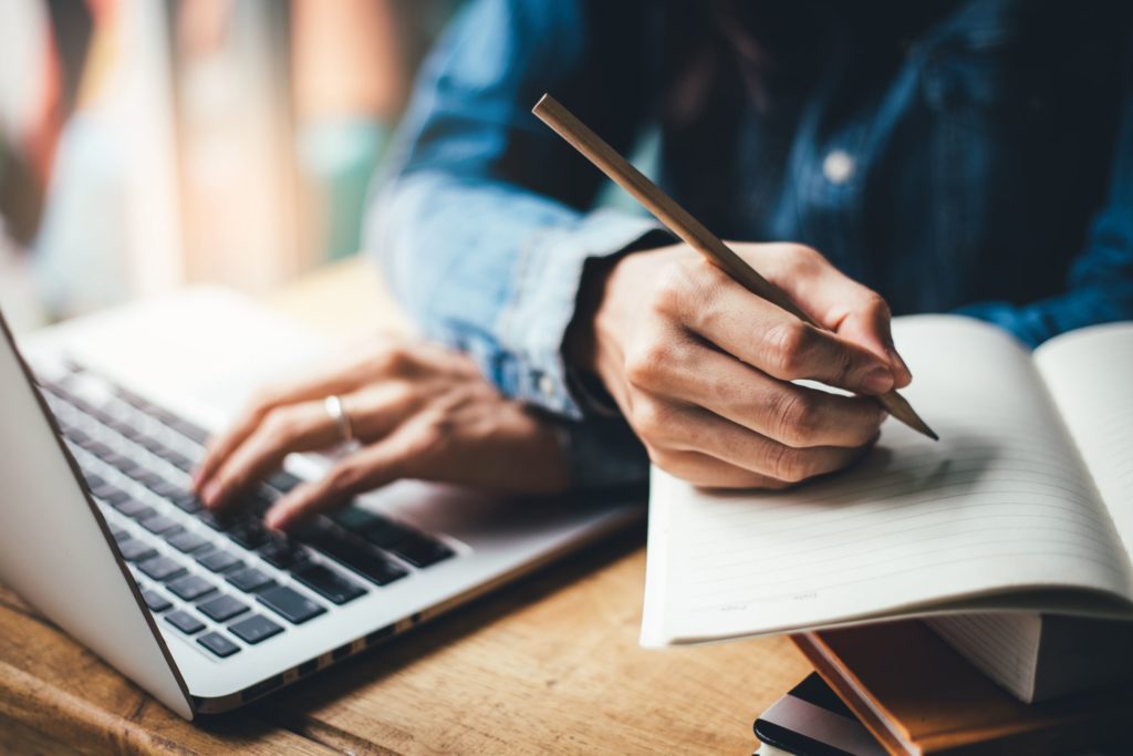 A close-up of a person’s hands as they research on a computer and write in a notebook.