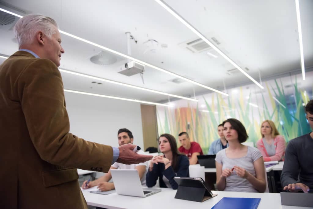 Older teacher lecturing to college students in a classroom.