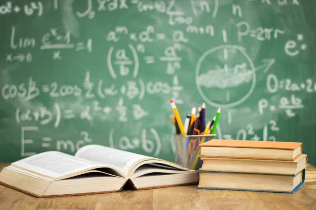 School books sit on a teacher’s desk in front of a chalkboard.