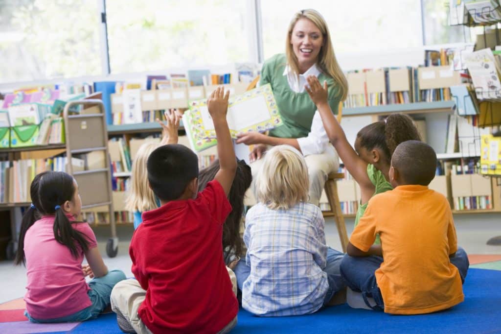 Teacher sitting and reading to a group of young students on the classroom floor.
